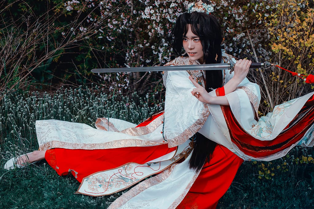 Ilia is cosplaying as Xie Lian wearing a layered red, gold, and white hanfu that is hand-painted with flowers. They are crouching and holding a sword in front of their chest in a defensive pose.