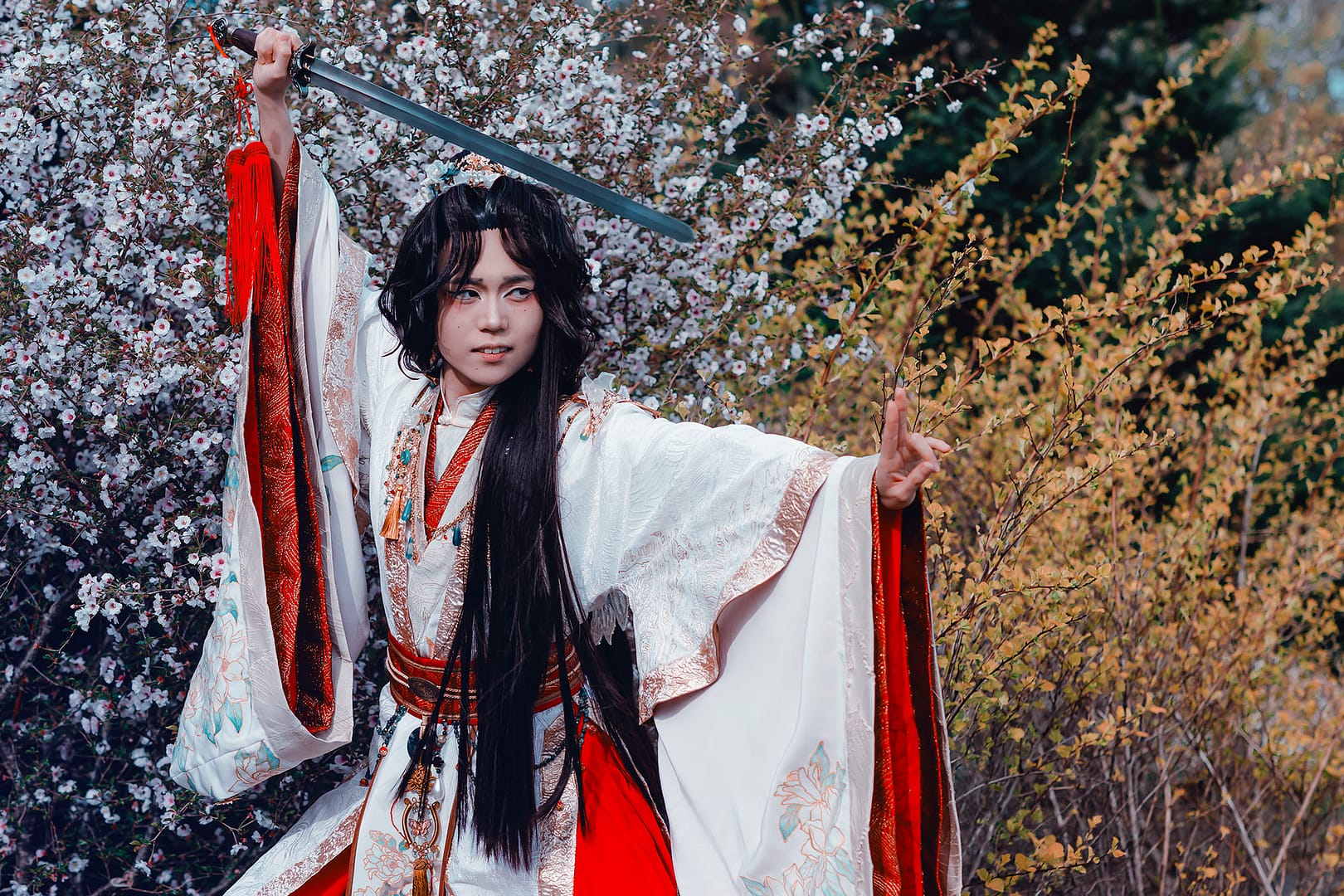Ilia is cosplaying as Xie Lian wearing a layered red, gold, and white hanfu that is hand-painted with flowers. They are holding a sword above their head prepared to strike.