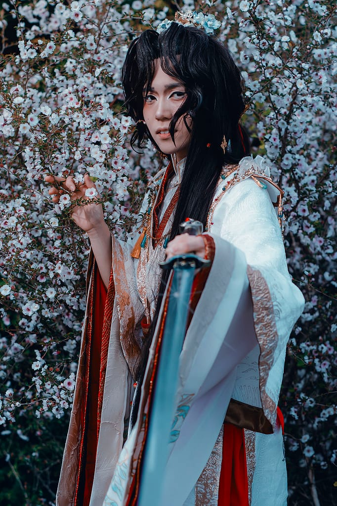 Ilia is cosplaying as Xie Lian wearing a layered red, gold, and white hanfu that is hand-painted with flowers. They are holding a branch of cherry blossoms in one hand and a sword pointed at the viewer in the other hand.