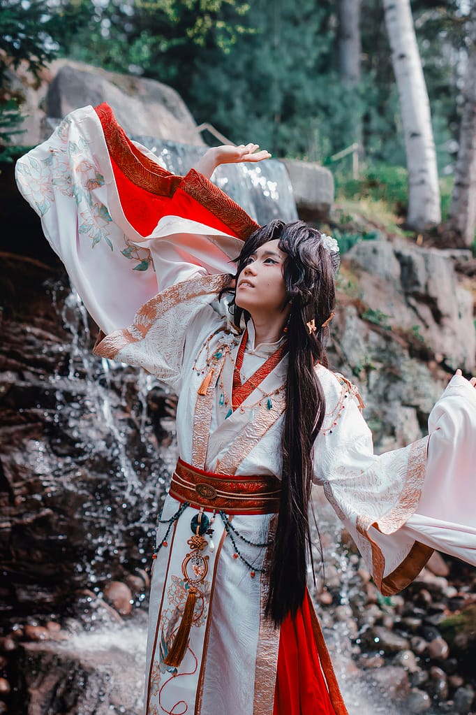 Ilia is cosplaying as Xie Lian wearing a layered red, gold, and white hanfu that is hand-painted with flowers. They are standing in front of a crashing waterfall and their sleeves are billowing in the wind.