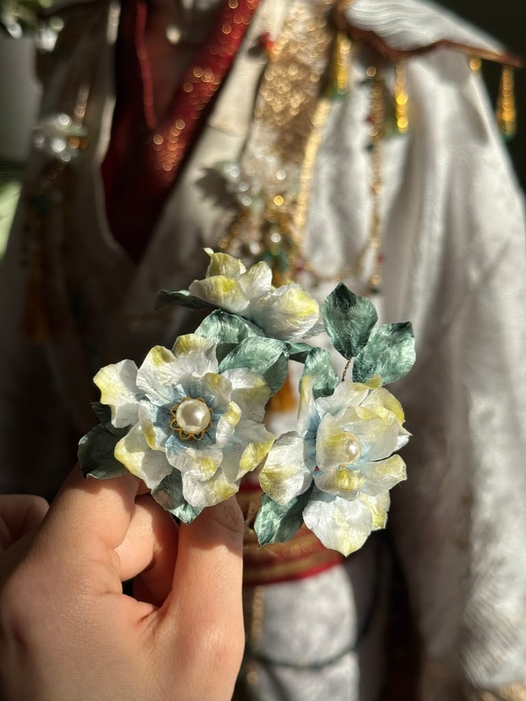 Ilia is holding their handmade white, blue, and yellow ronghua (silk velvet flowers).